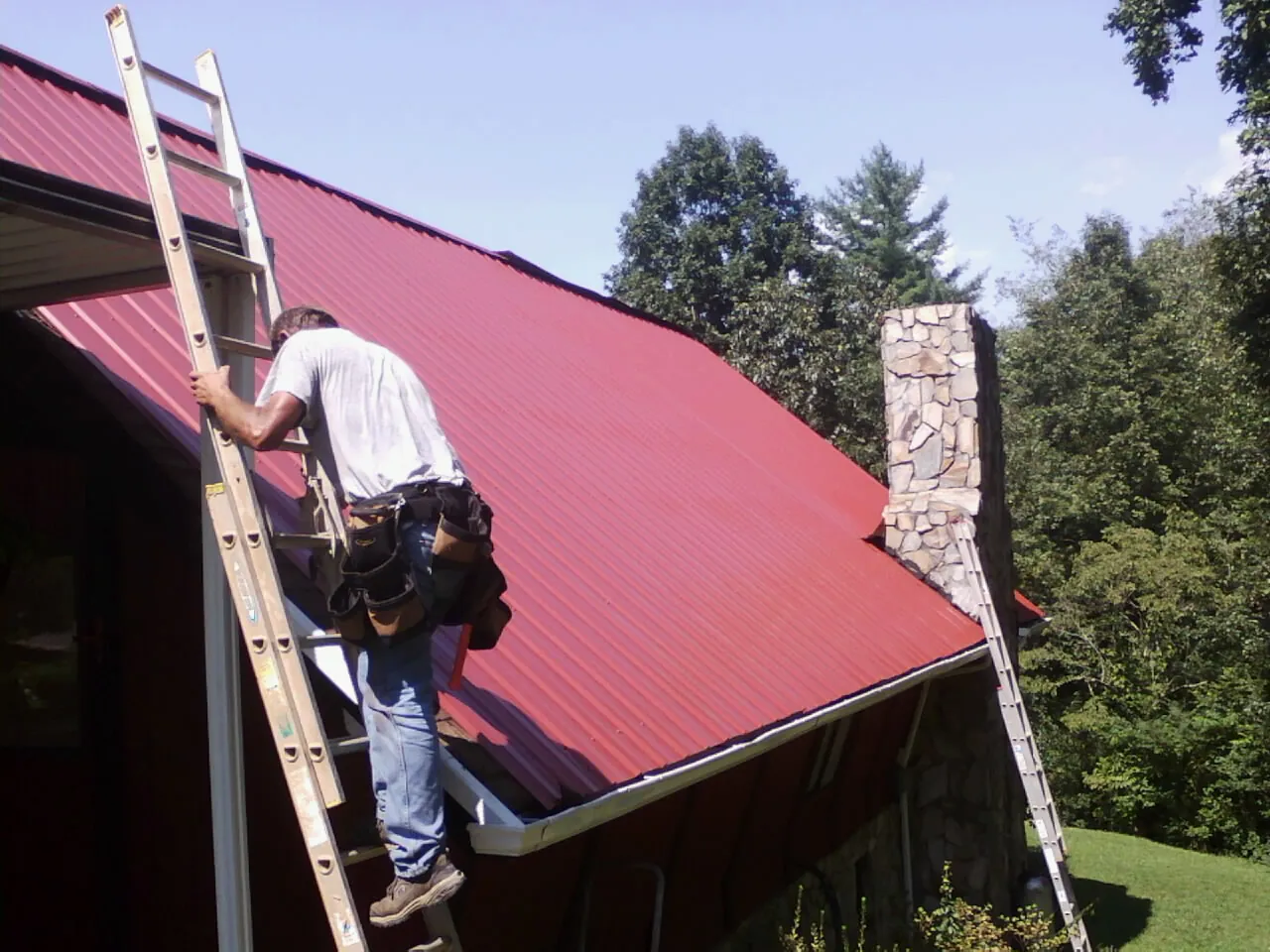 Roofer on a ladder beside a red metal roof and stone chimney — Soffit & Fascia Repair by Roofing Brothers in Passaic